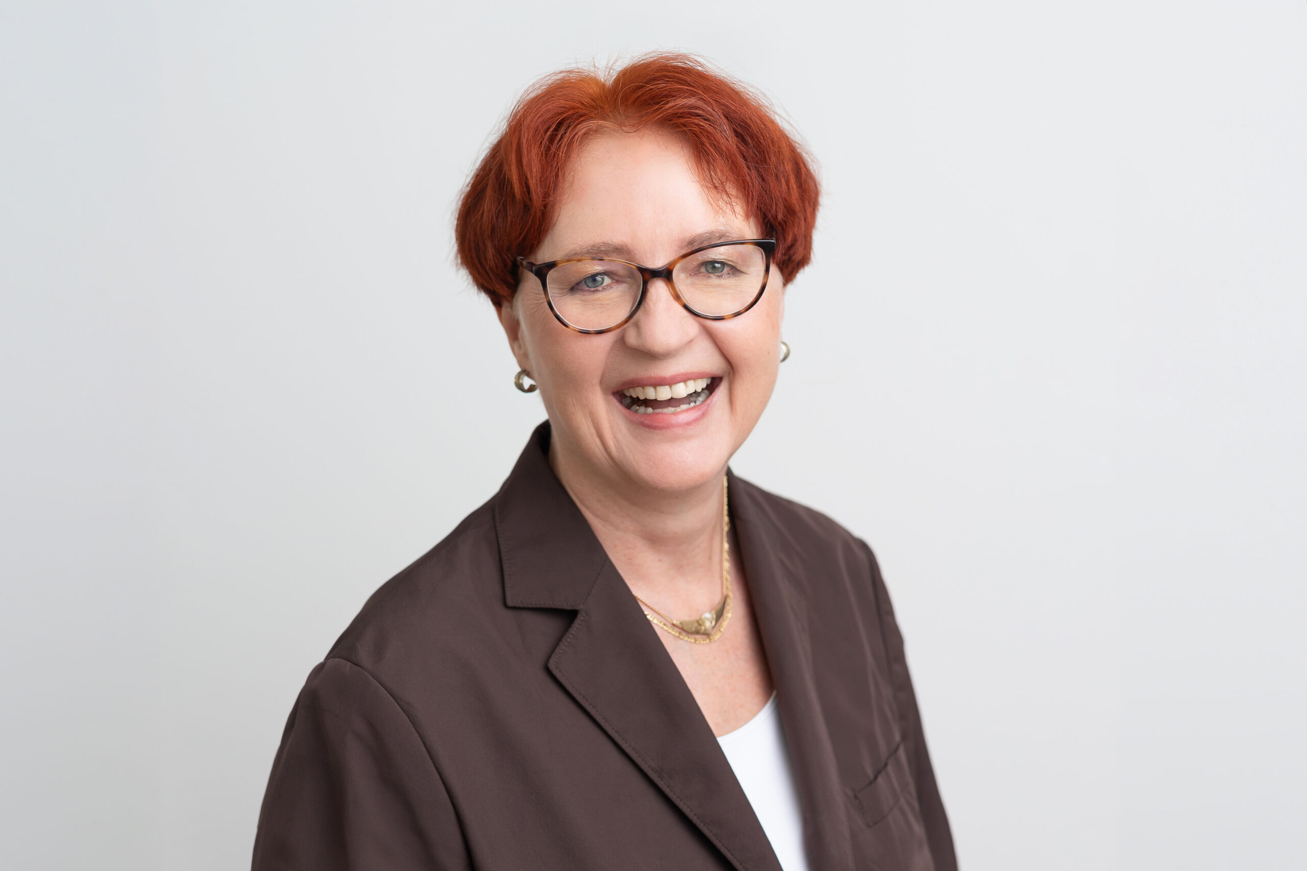 Woman with red hair and glasses in brown blazer, smiling headshot.